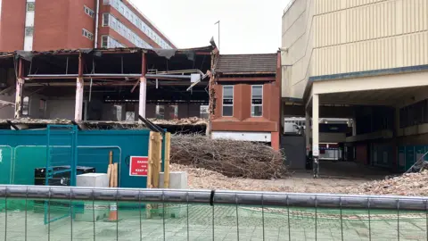 David Freezer/BBC Wooden hoardings and metal fencing stand before partly demolished two-storey shops at the front of a shopping complex, with bricks and insulation material piled on parts of the pavement.