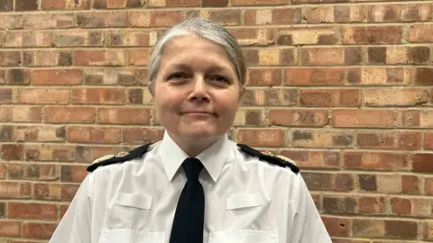 Standing in front of a bare brick wall, Sarah Crew smiles at the camera. She wears a white Avon and Somerset Police shirt and a black tie. 