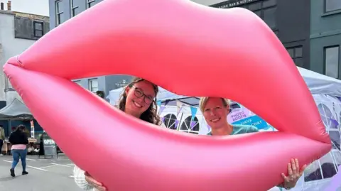 Bright Tights A pink pair of giant inflatable open lips dominate the image, with the heads of two smiling women visible from within the mouth.