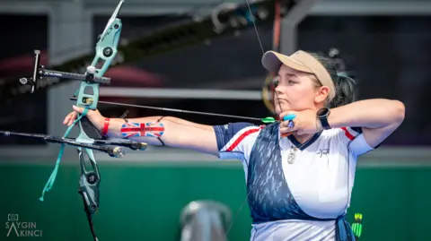 A woman with a white and blue top, tied-back brown hair and a light brown cap holding a bow and arrow and looking down a sight