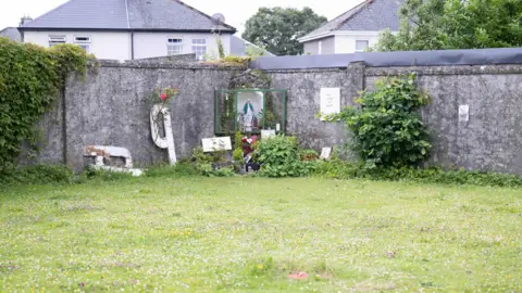 Andrew Downes/ODAIT/PA Wire A grassed courtyard encased by a plain grey wall, in which a Catholic shrine has been erected in one corner. The shrine contains a statue of Our Lady in blue and white robes. The pitched roofs of semi-detached houses can be seen on the other side of the walls. 