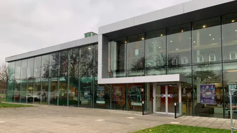 Shariqua Ahmed/BBC The Key Theatre - a glass-fronted building with paving and grass in front of it. It is two-storeys. It is a cloudy day.