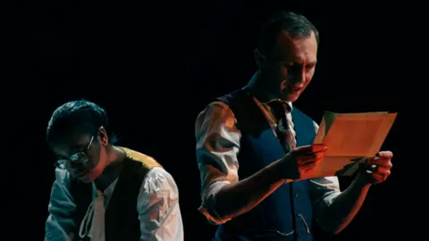 Neil Reading A woman with glasses and short black hair is wearing 1950s-style clothing and looking down while a man in a navy waistcoat is standing tall and looking at sheets of paper, as both are in front of a black background.