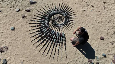 Overhead shot of a man crouched over a spiral formation of rocks and pebbles on a beach