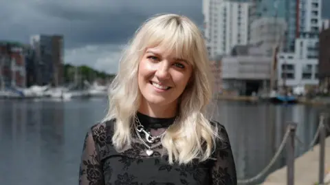 A woman with blonde hair stood by a large body of water wearing a black top