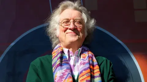 Noddy Holder - a man with long grey hair, wearing glasses, a colourful scarf, purple and white shirt, and green jacket - stands and smiles.