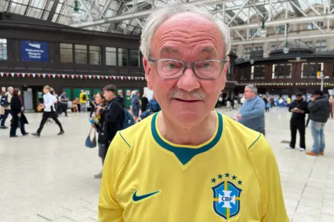 BBC Malcolm McLean, an older man wearing glasses and a bright yellow Brazil national football jersey stands in the foreground of a large indoor train station concourse. He is facing the camera. The spacious hall features a high glass-and-steel roof, dark wooden buildings along the sides, and people walking in the background. A sign reading “Welcome to Glasgow Central” is visible behind him.