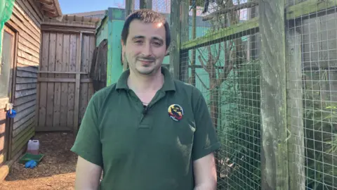 BBC/Emily Doughty Connor Gordon is wearing a green shirt. He is standing next to a cage containing marmosets, with a wooden frame and wire mesh. There are tall wooden fences behind him and wooden buildings.