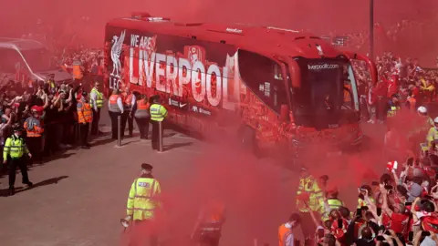 Reuters Liverpool-branded coach with the squad drives through the red mist from flares, with fans crowding on the pavements and police officers with high-visibility coats postioned around