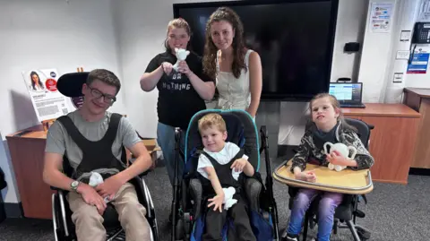 Four children and a designer in a classrom room. Three children are in wheelchairs, one is stood at the back with the woman, who designed Moti Me.