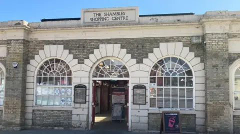 The entrance to 'The Shambles Shopping Centre' with a sign outside for the Twisted Mallow sweet shop. Three white stone arches are on the front of the building.