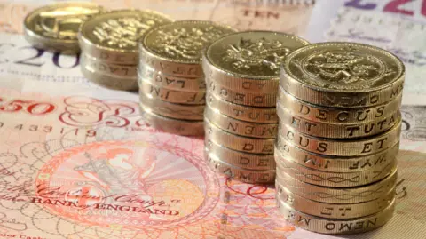 Piles of pound coins are placed, growing in size as they come closer to the camera. They are sitting on notes including £50 and £20 which are just seen.