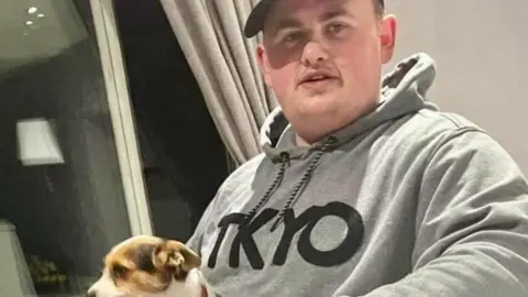 Bedfordshire Police Man smiling to camera. He is wearing a cap and grey hooded top. He has a small Jack Russell puppy, brown, black and white, in his lap.