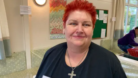 Volunteer Heather Windows with very short red hair wearing a cross and a dark top in a hall smiling at the camera