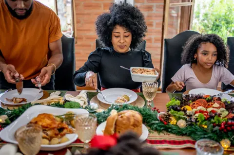 Getty Images Family around the table enjoying a Christmas dinner. A woman serves herself potatoes in the middle of the frame, next to a man and a young girl, with a selection of festive food and decorations all over the table in the foreground.