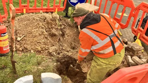 BBC An engineer digs a hole in the ground with a shovel. He wears an orange hi-vis jumper, knitted hat and trousers. Orange temporary fencing surrounds him.