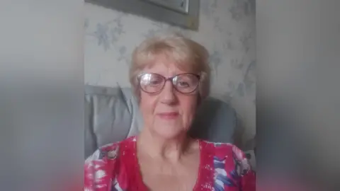 A selfie of a woman with short blonde hair, she's wearing a pink t-shirt with blue and white flowers on it and purple framed glasses. She is sitting on a light blue chair, which can be seen behind her alongside white wallpaper with light blue flowers and the corner of a picture frame. 