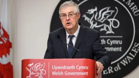 Getty Images Mark Drakeford Berdiri di Pemerintah Welsh bermerek Lecturn yang menyelenggarakan konferensi pers selama pandemi. Dia memiliki tanda bilingual abu -abu besar yang terbuat dari batu tulis di belakangnya mengatakan pemerintah Welsh dan menteri pertama Wales dan bendera Welsh di sebelah kanannya