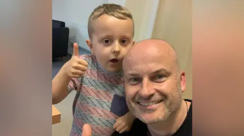 Kev Pitcher Kev Pitcher kneels and his son Benny rests an arm on his shoulder. Both are smiling and have their thumbs up. 