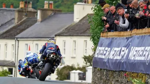 O IOM tt ande a três motos de TT, uma fileira de casas na frente delas e os fãs estão encostados na parede de pedra à direita.