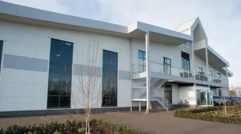 David Lloyd A large white building with tall tinted-glass windows and a large arch above the entrance and small shrubs on a paved area in the foreground