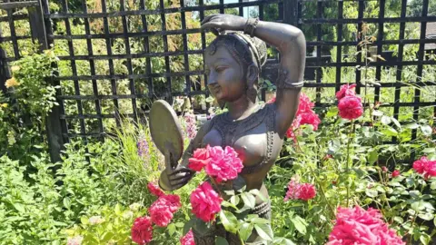 A bronze statue of an Indian woman is placed among pink flowers in a garden. She is designed as if wearing ornate clothing and looking at herself in a mirror. There are shrubs behind her and a trellis fence.