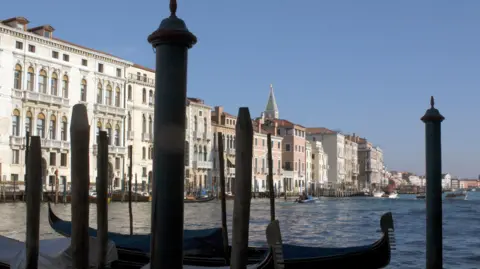 A waterfront in Venice with wooden posts and gondolas in the foreground and a row of buildings and more boats in the background.