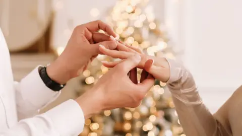 A person's hands put a ring on to another person's hand in front of a Christmas tree