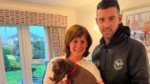 Julie Fripp holding Frippy, who seems to be licking her hand, with them standing next to Dave Rimmer in what appears to be a living room.