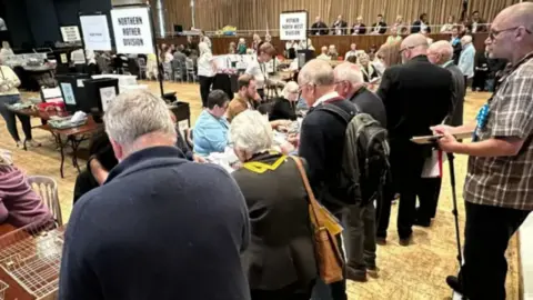 A crowded indoor hall shows people standing in a long line, facing rows of tables staffed by officials. Signs are visible above the tables, and the space resembles a community voting or counting venue.