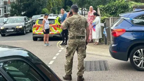 SWNS A soldier stands on the phone with his back to camera as people look on at the scene in Sally Port Gardens, Gillingham