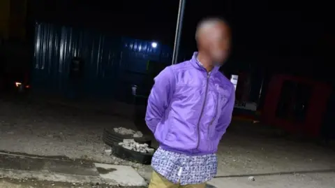 A man, with his face blurred, is pictured at night time wearing a purple jacket and khaki trousers.