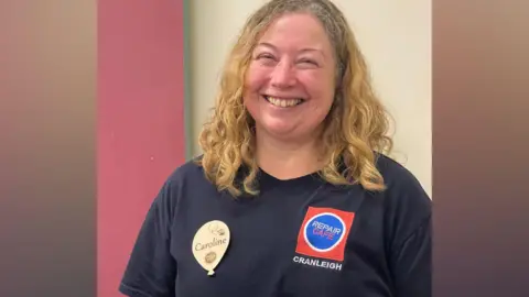 A blonde woman wearing a dark navy shirt, with a logo that reads Repair Cafe Cranleigh. She is also wearing a badge that reads Caroline.