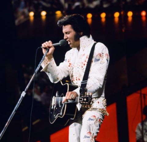 Elvis Presley sings into a microphone. He is wearing a white sparkly suit and playing a black acoustic guitar