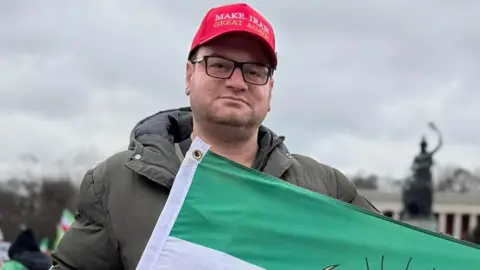 Pasha Pour Pasha Pashapour Alamdari. He is pictured holding an Iranian flag - only the green and white stripe can be seen. He is wearing a red cap that says "Make Iran great again"