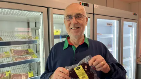 Ellen Knight/BBC Grahame Hurst stans in front of the farm shops fridges, which are full of packets of bacon and black pudding. He is smiling and has short white hair, and is wearing a pair of thin-framed glasses. He is wearing a dark blue rugby shirt with a green collar. 