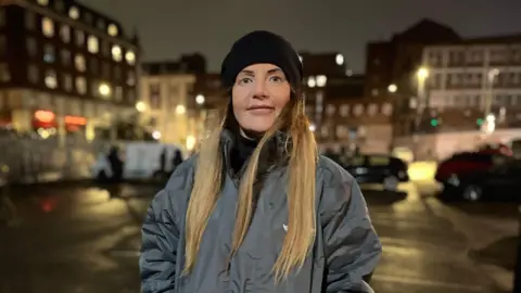 Becky Joyce looks directly into the camera. She is wearing a grey coat and black hat. Out of focus behind her are a car park and buildings in the centre of Leeds