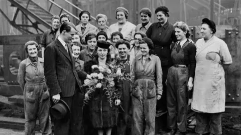 A black and white photo with about two dozen women wearing various shipbuilding uniforms standing in three rows. At the front, a woman in a hat and fur coat is being presented with a large bouquet of flowers . A man in a pinstripe suit stands beside her.