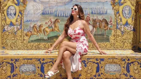 A woman wearing a white dress with a red flower pattern, white sandals and white-framed sunglasses sits against an ornately tiled backdrop.
