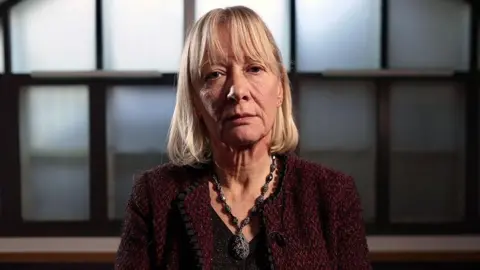 A portrait of lawyer Sally Marsden. She is seated in front of an arched window and has golden hair. She is wearing a statement necklace, black top and maroon jacket.