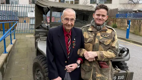 BBC/Elliott Green Richard on the right, in army camo gear, links arms with Arnold, on the left. They lean against the bonnet of a dark green truck with large tires and a fabric roof.