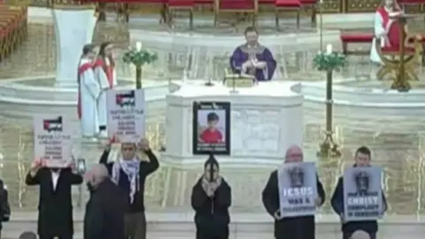 Protesters holding up placards with the Palestinian flag on them standing at the front of a cathedral.  A priest in purple robs is standing behind an alter behind them.