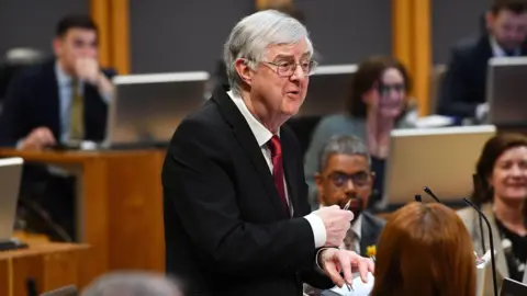 PA Media Mark Drakeford addressing the Senedd surrounded by politicians from the Welsh parliament. He's wearing a black suit and red tie.