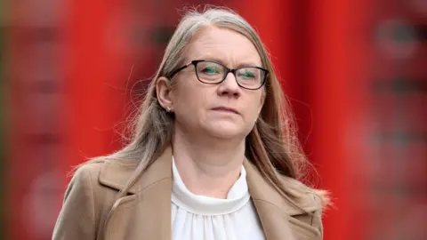 Shirley Anne Somerville walking along a street. She has long blonde hair and is wearing dark rimmed glasses. She is wearing a beige coat and a white blouse.