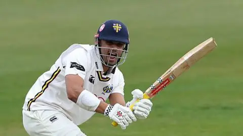 Left-hander Emilio Gay, wearing his navy blue Durham helmet, watches after playing a shot into the legside with his bat in the air 