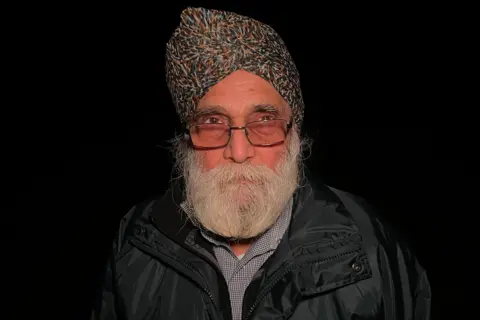 Sam Read/BBC Surinder Bhoday is wearing a turban and glasses. He has a white beard. He is wearing a coat and shirt. It is nighttime.