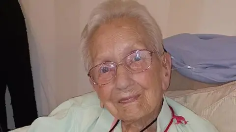 A close-up photo of an elderly woman with short grey hair, blue eyes and glasses. She is wearing a pale green shirt.