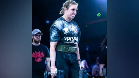 Pursuit Images A woman during a strongwoman competition carries a bar in each hand. She is grimacing as she lifts. She has dark hair that has been plaited back. She wears a black T-shirt covered in white chalk, black leggings and a supportive belt. 