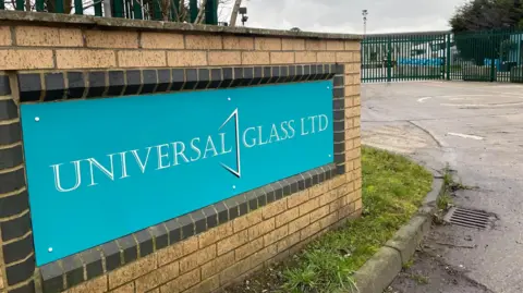 Simon Thake The entrance to an industrial site with a green sign fastened on to a brick wall with the words, 'Universal Glass'