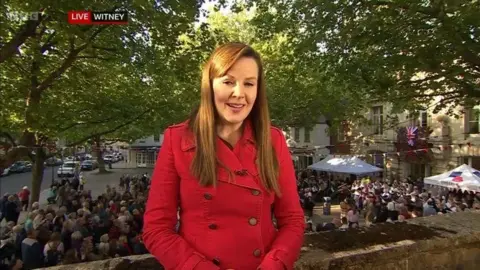 Alexis Green stands in front of crowds of people attending a street party to report on the weather. She has long light brown hair and wears a red trench coat.